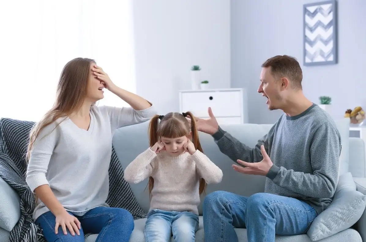 Ein Kind sitzt zwischen zwei Erwachsenen auf einem Sofa und hält sich die Ohren zu.