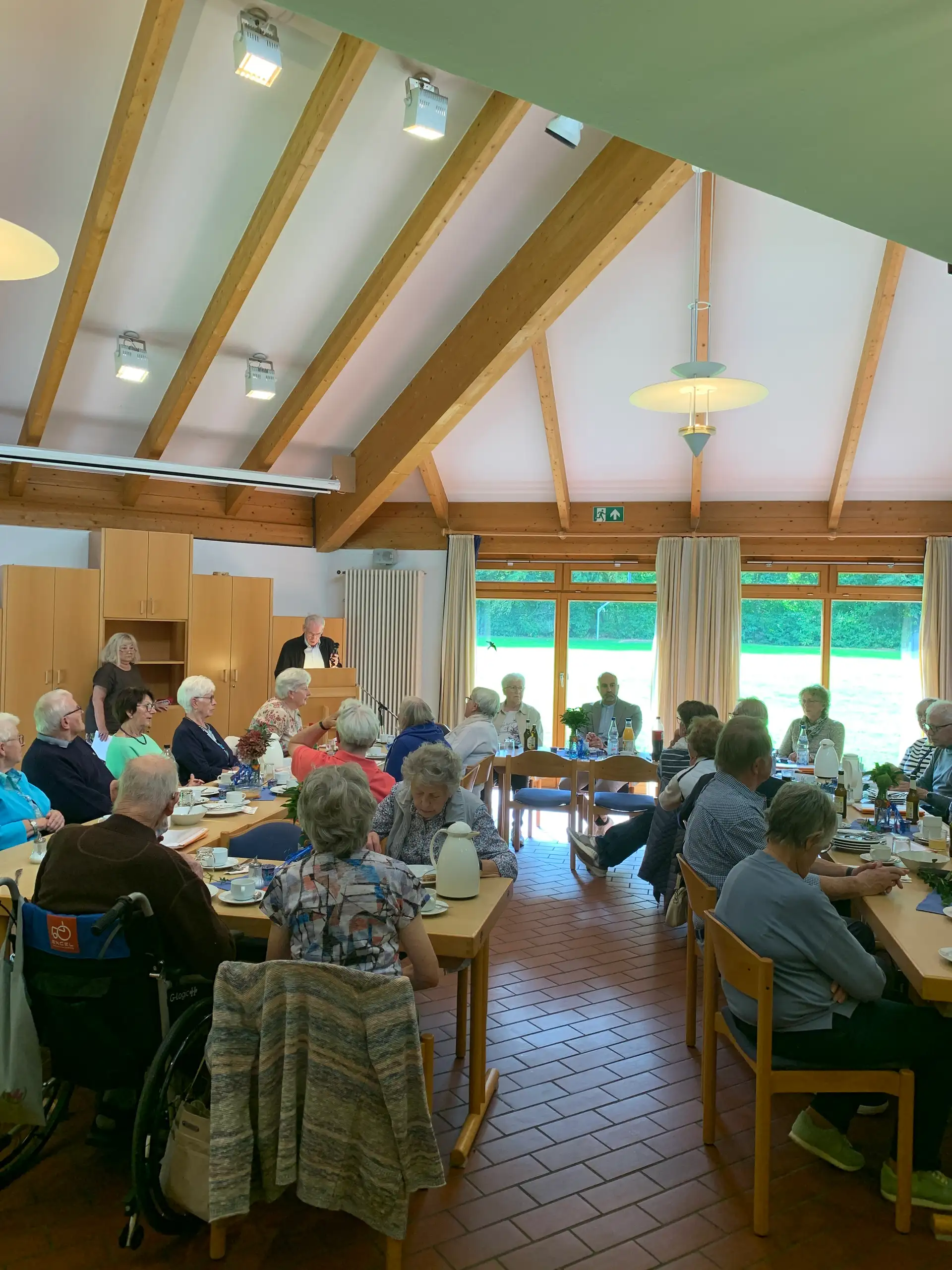 Menschen sitzen bei einem Treffen im Gemeindehaus an Tischen; eine Person spricht vorne am Pult.