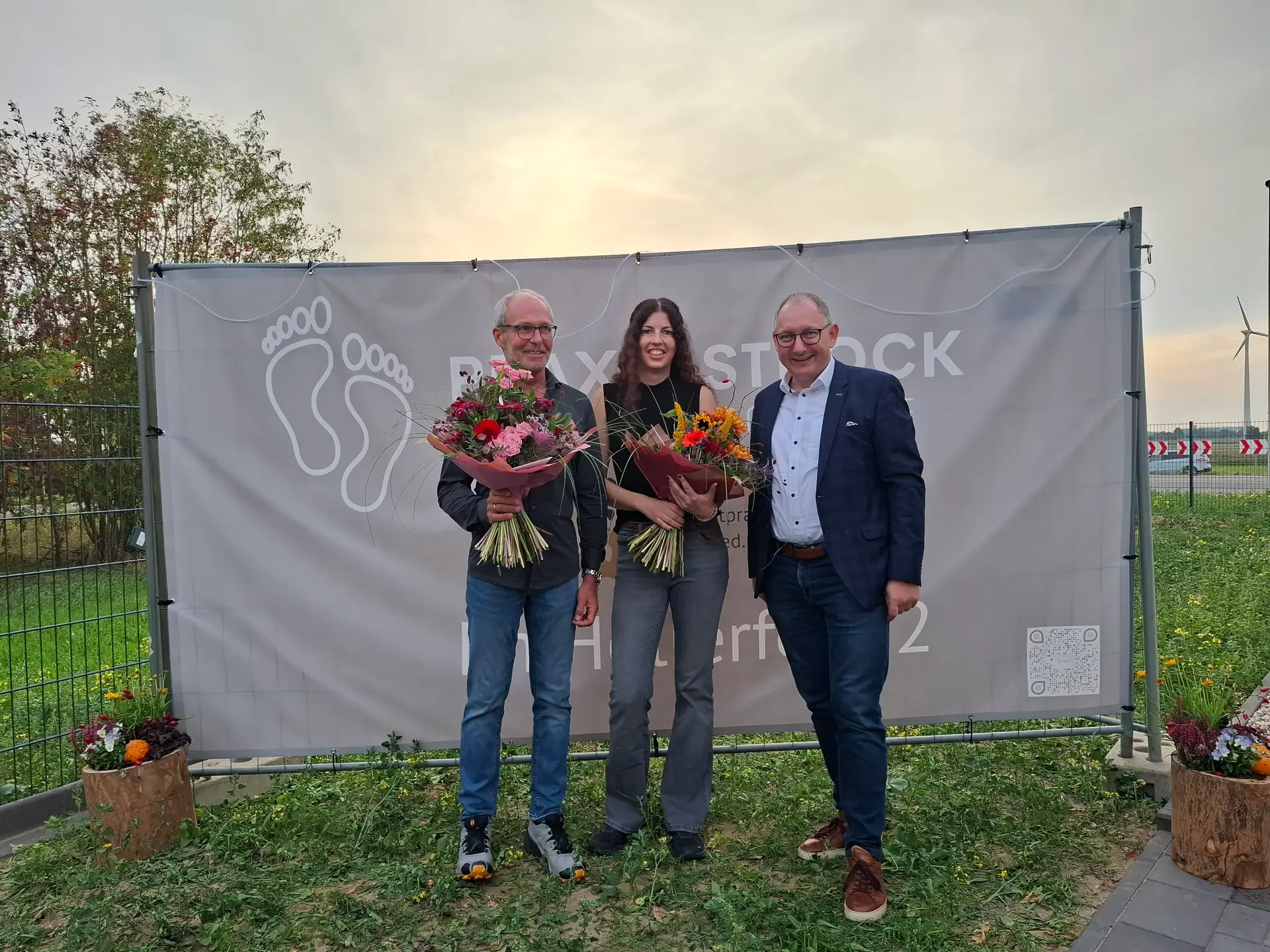Drei Personen stehen vor einem Praxis-Strock-Banner im Freien; zwei von ihnen halten Blumensträuße.