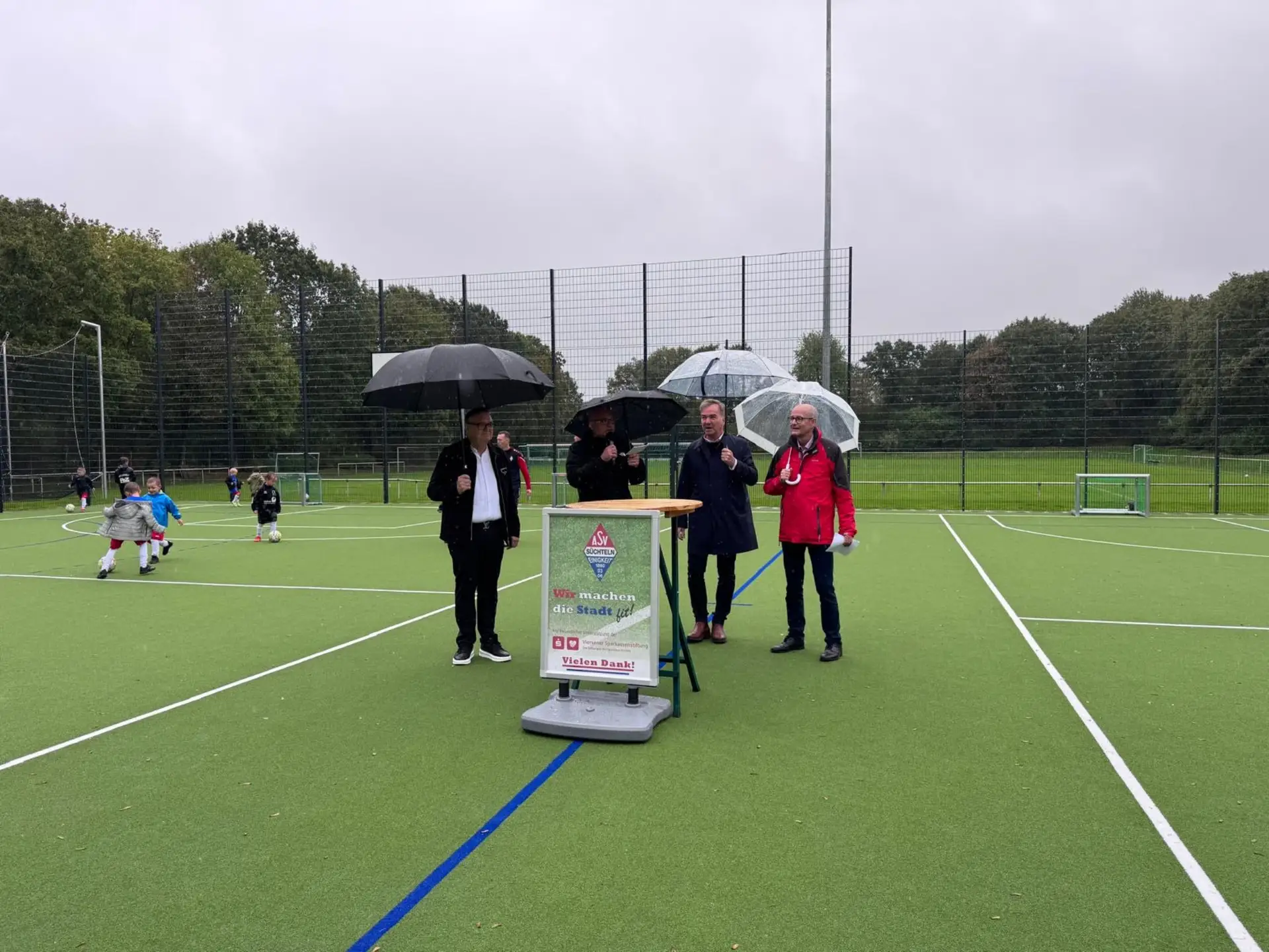 Personengruppe bei der Eröffnung eines Multifunktionsspielfelds, im Hintergrund spielen Kinder auf dem neuen Sportplatz.