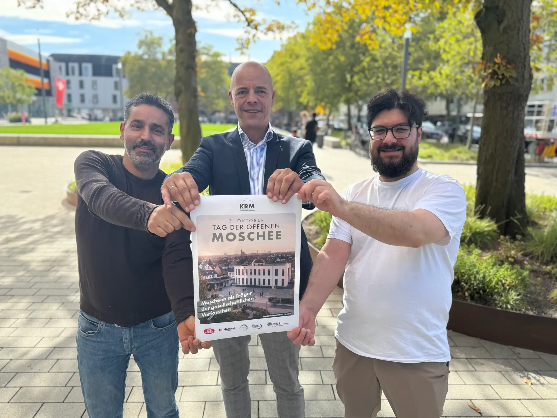 Drei Personen stehen nebeneinander und blicken in die Kamera; im Bildzusammenhang wird ein Besuchsangebot zum Tag der offenen Moschee angekündigt.